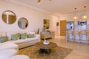 Ein modernes Wohnzimmer mit einem weißen Sofa, einem Couchtisch und einer Küchenzeile inklusive Kühlschrank und Barhocker.