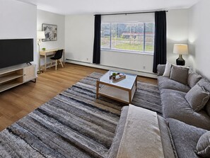 Ein modernes Wohnzimmer mit einem Flachbildfernseher, einem Sofa, einem Couchtisch und einem großen Fenster mit Jalousien.