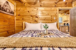 Ein Holzhütten-Innenraum mit einem Bett, Kissen, einer Steudecke, einem gewebten Teppich und einem Bild an der Wand.