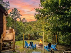 A wooden deck with a fire pit and blue chairs, surrounded by lush greenery and a forested backdrop. - Starlit Ridge – Hot Tub, Fire Pit, Mountain Views (Sevierville)