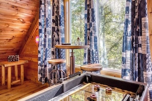 A cozy cabin interior with a wooden sloped ceiling, a rustic coffee table, a small wooden stool, and a window with patterned curtains. - Starlit Ridge – Hot Tub, Fire Pit, Mountain Views (Sevierville)