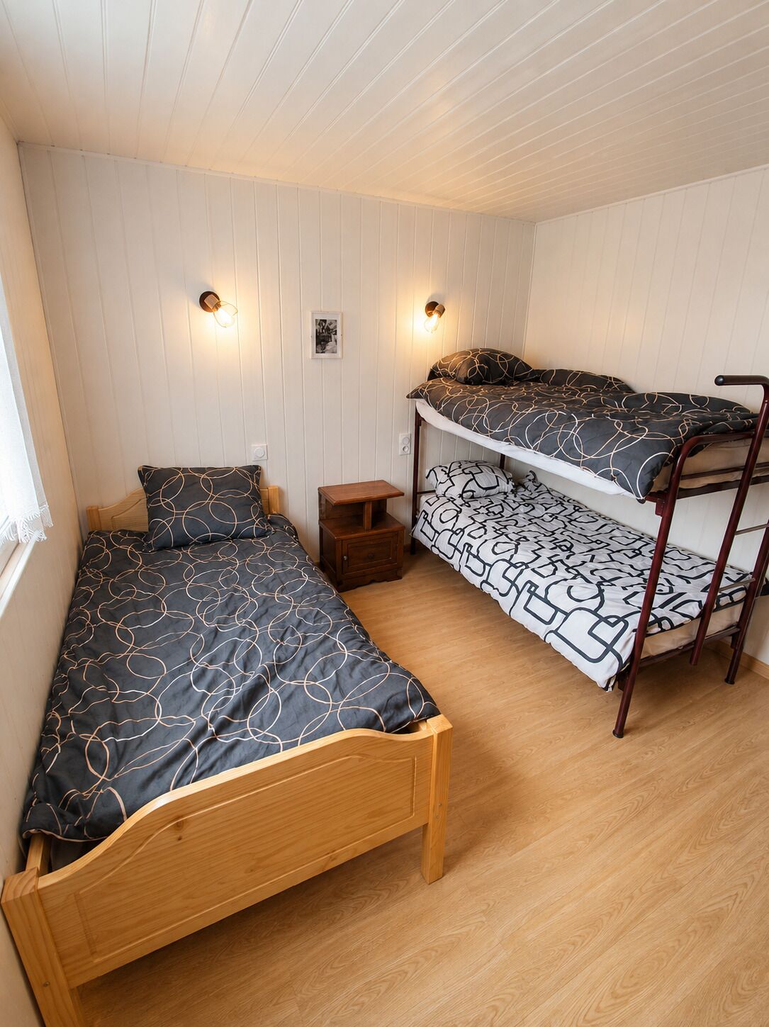 Una habitación con dos literas, una cama individual, un pequeño mesita de noche y dos lámparas fijadas a la pared.