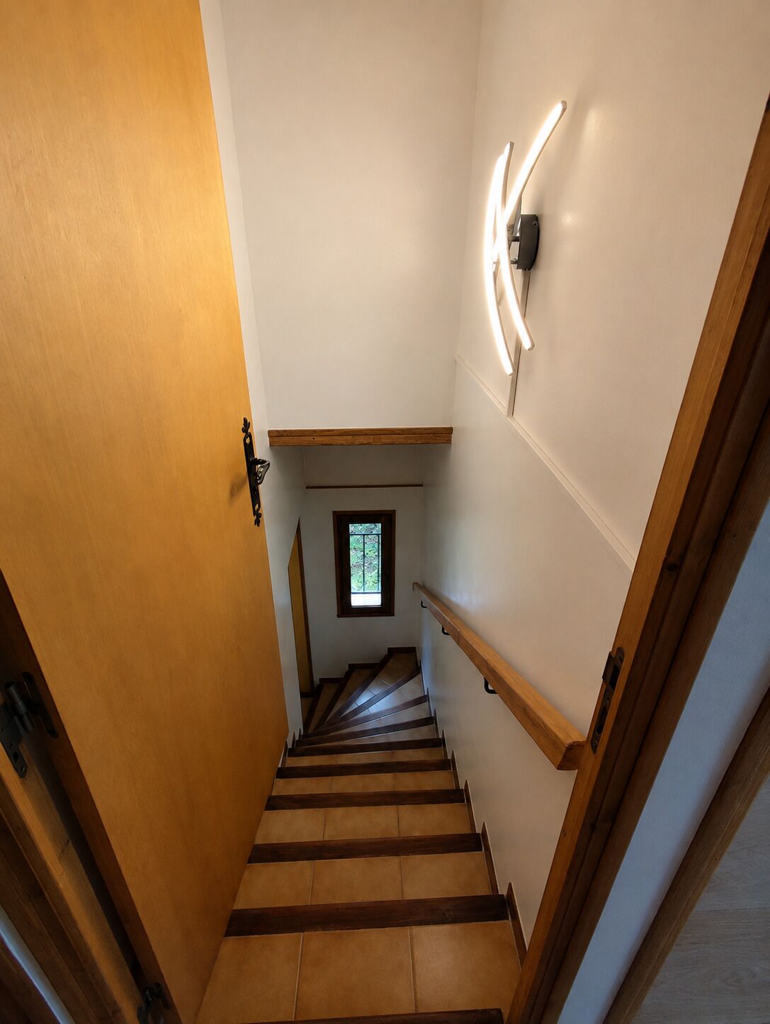 Una escalera de madera con barandilla, un pequeño ventanal en la parte superior e iluminación empotrada en la pared.