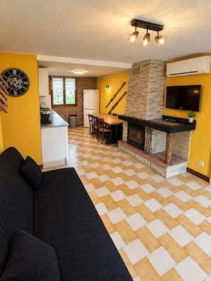 Un espacio habitable moderno con piso a cuadros, un sofá, una zona de comedor, una cocina con chimenea de piedra y un televisor montado.