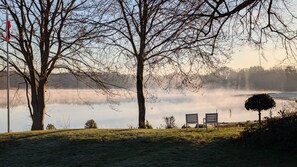 Eine friedliche Seenlandschaft mit zwei Bänken, die zum Wasser hin ausgerichtet sind, umgeben von kahlen Bäumen und einem klaren Himmel.