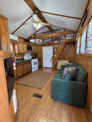 A cabin interior with a kitchenette, a green sofa, wooden beams, and a staircase.