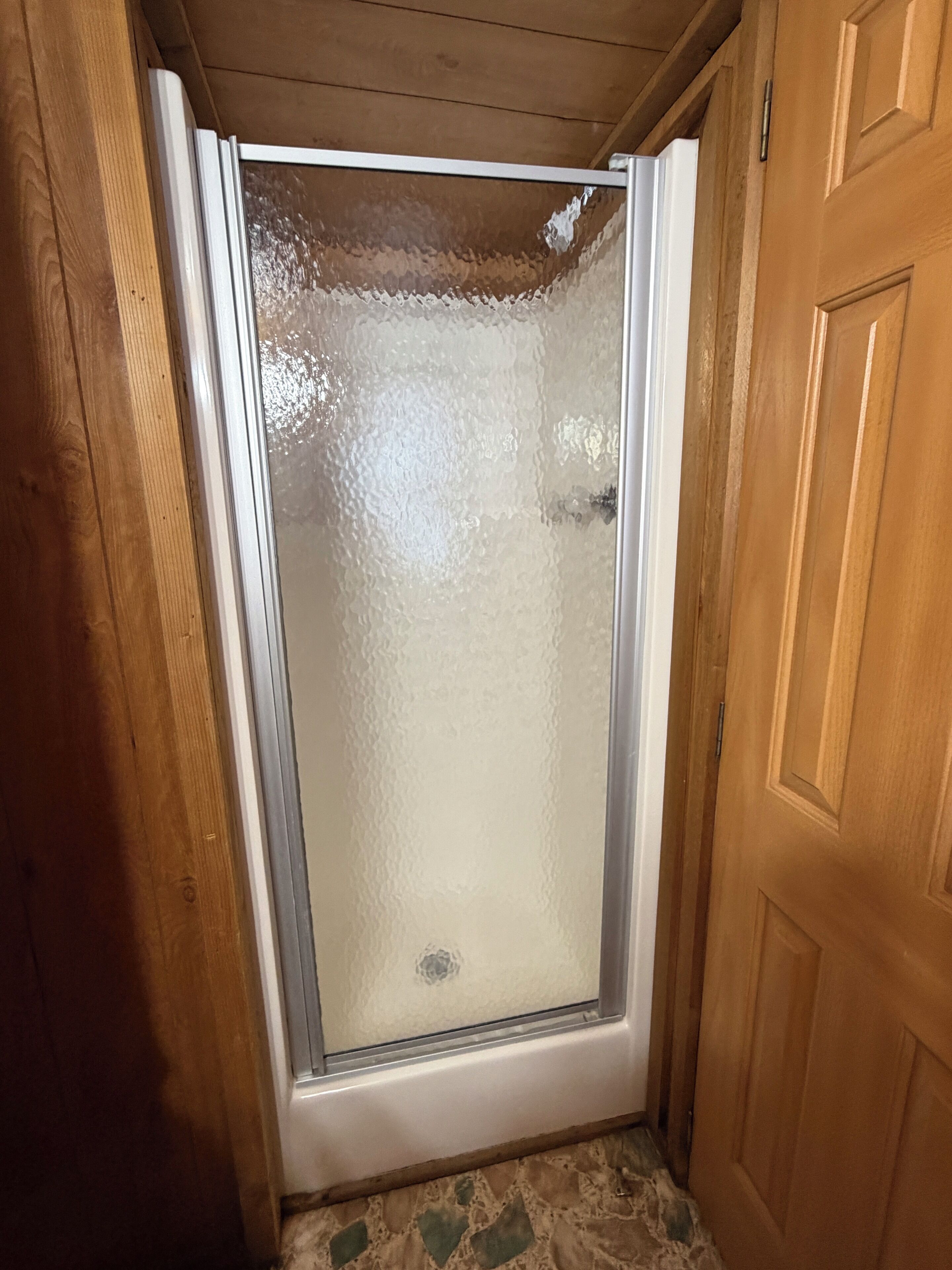 Una doccia con porta in vetro acidato, pareti in legno e pavimento piastrellato.