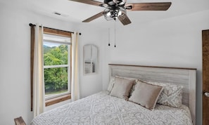 Een slaapkamer met een bed, een plafondventilator, een raam met gordijnen en uitzicht op groen.