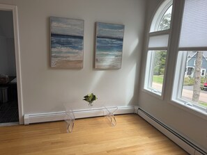 Ein Zimmer mit zwei Bildern, einem kleinen Tisch mit einer Vase und einem großen Fenster.