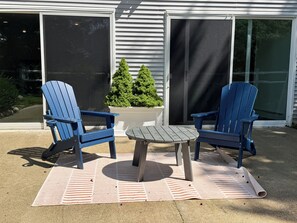 Zwei blaue Adirondack-Stühle und ein kleiner Tisch auf einer gemusterten Terrassenmatte, mit einem Blumentopf und einem Gebäude im Hintergrund.