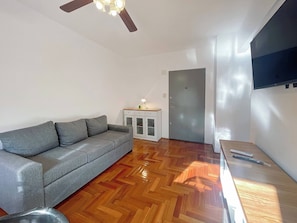 Een kamer met een grijze bank, houten visgraatvloer, een plafondventilator, een kast met lades, een deur en een flatscreen televisie die aan de muur is bevestigd.