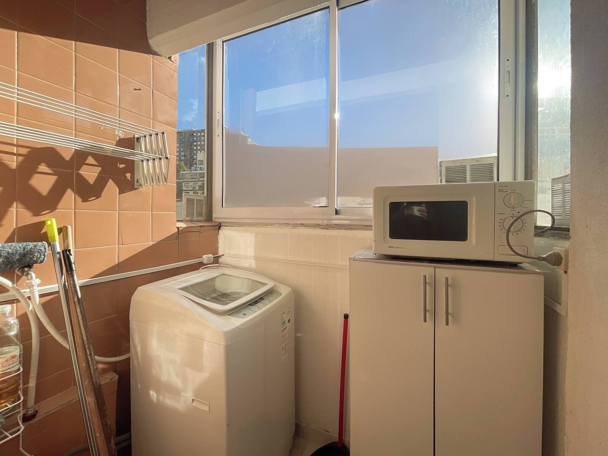 A compact kitchen with a microwave on top of a cabinet, a washing machine, and a window with a view of buildings.