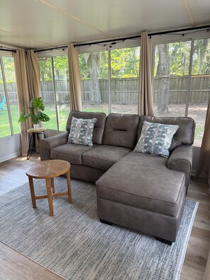A modern living room with a sectional sofa, a small wooden table, and a large window with curtains. - The Palmetto Blue Cottage Just Minutes to Parris Island! (Port Royal)