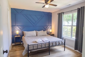 A bedroom with a bed, two bedside lamps, a ceiling fan, a window with curtains, and a wooden floor.