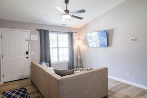 A living room with a sofa, a ceiling fan, a TV mounted on the wall, and a window with curtains. - The Palmetto Blue Cottage Just Minutes to Parris Island! (Port Royal)