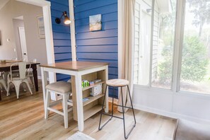 A room with a wooden table, a stool, a shelf with books, and a window with a view of greenery.