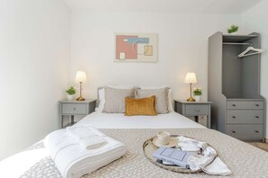 Una cama bien tendida con almohadas, una mesita de noche con lámpara, una bandeja con toallas y un libro, y un armario con perchas.