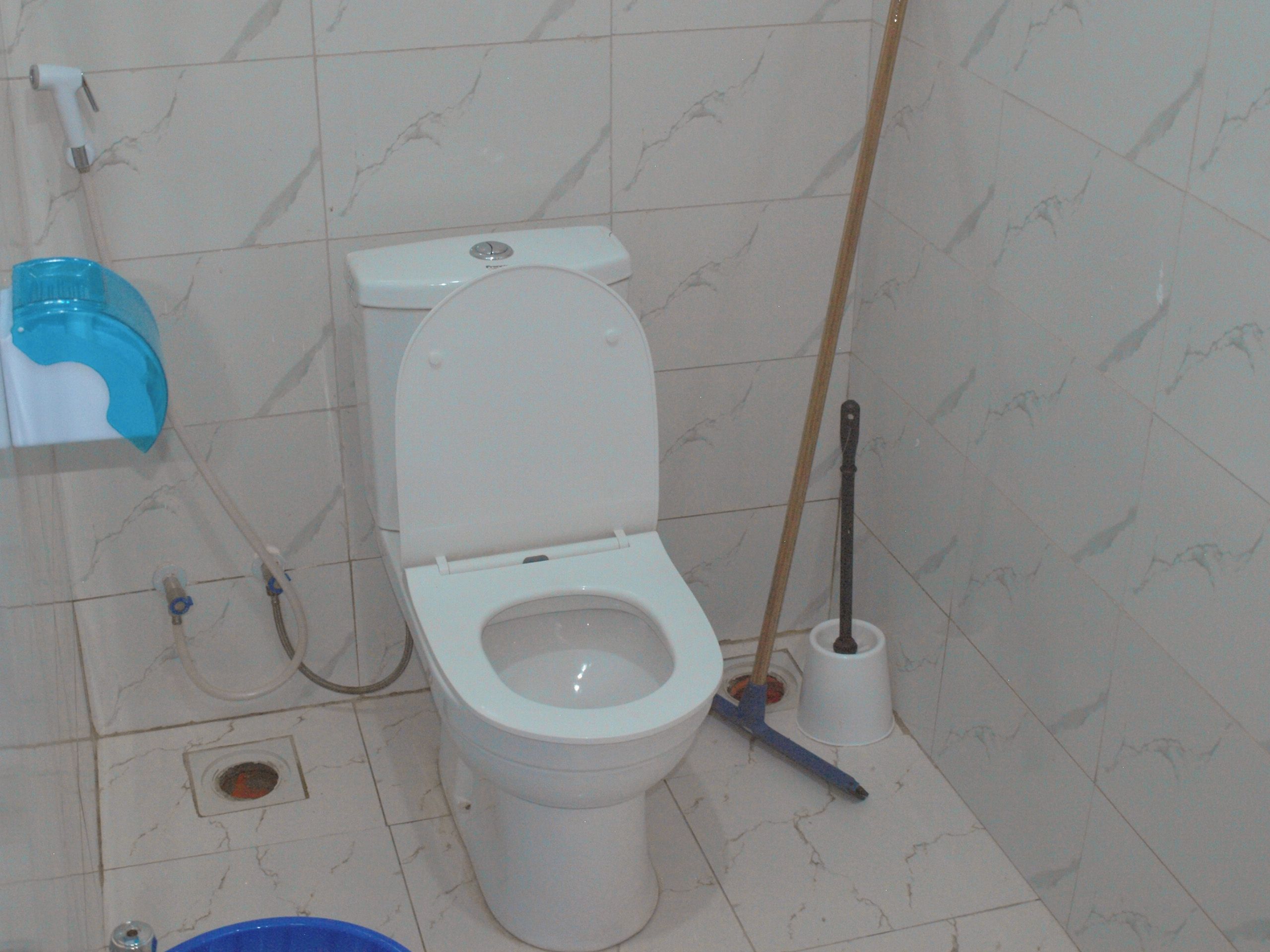 Ein Badezimmer mit Toilette, einem blauen Reinigungseimer, einem Pömpel und einer Toilettenbürste.