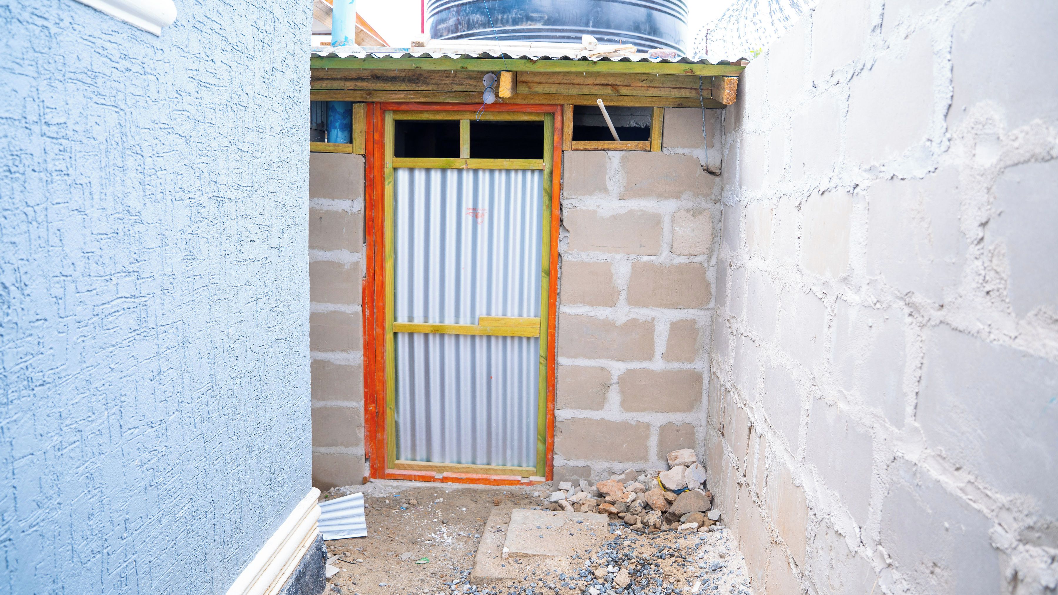Un viottolo stretto con una porta in lamiera ondulata, una piccola struttura in legno sopra e un muro di mattoni alla destra.