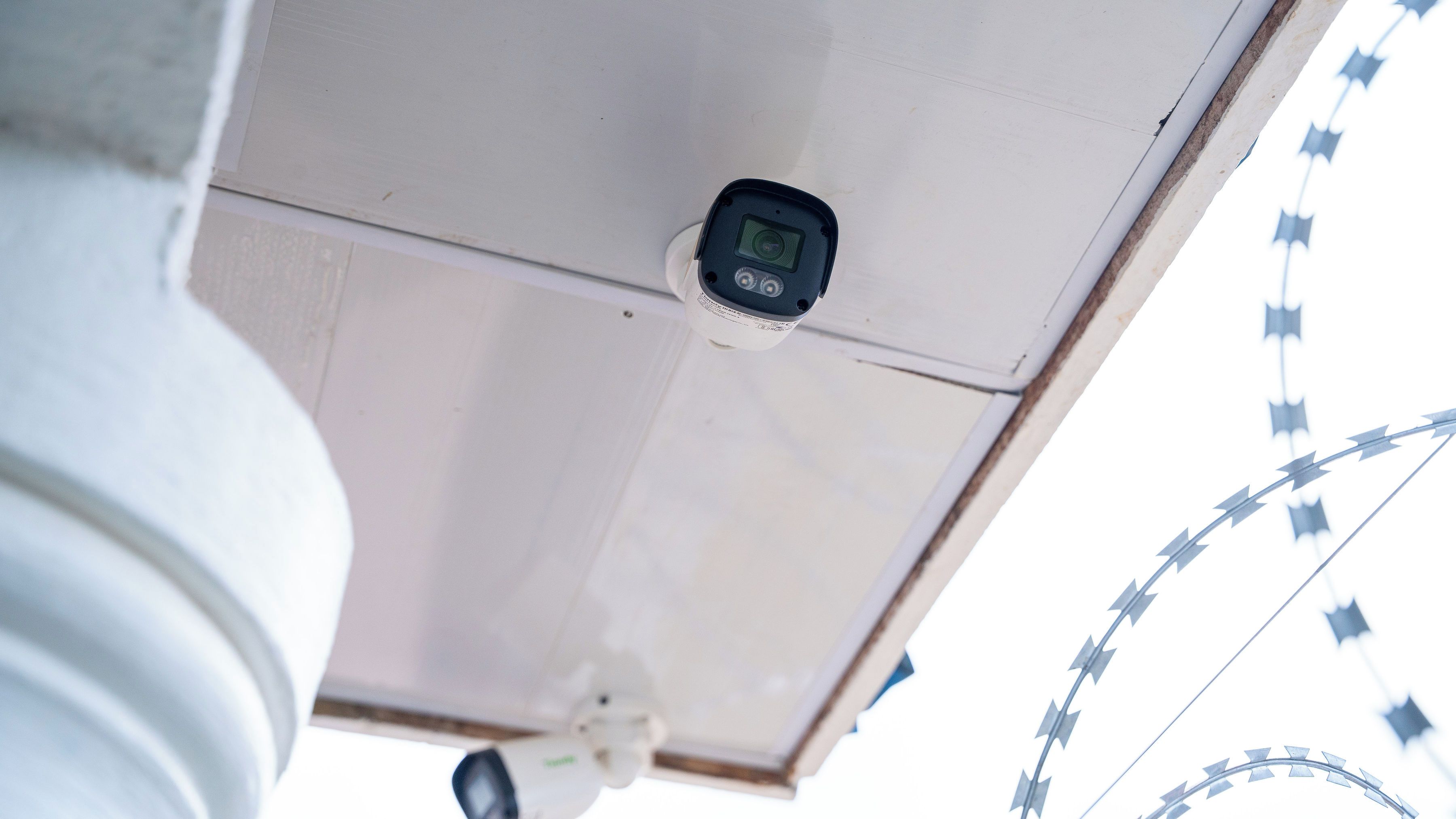 A security camera mounted on a ceiling, a white pipe, and a barbed wire fence in the background.