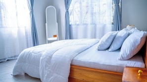 Ein ordentlich bezogenes Bett mit weißer Bettwäsche in einem Zimmer, das ein Holz-Kopfteil, einen Nachttisch und ein Fenster mit Vorhängen hat.