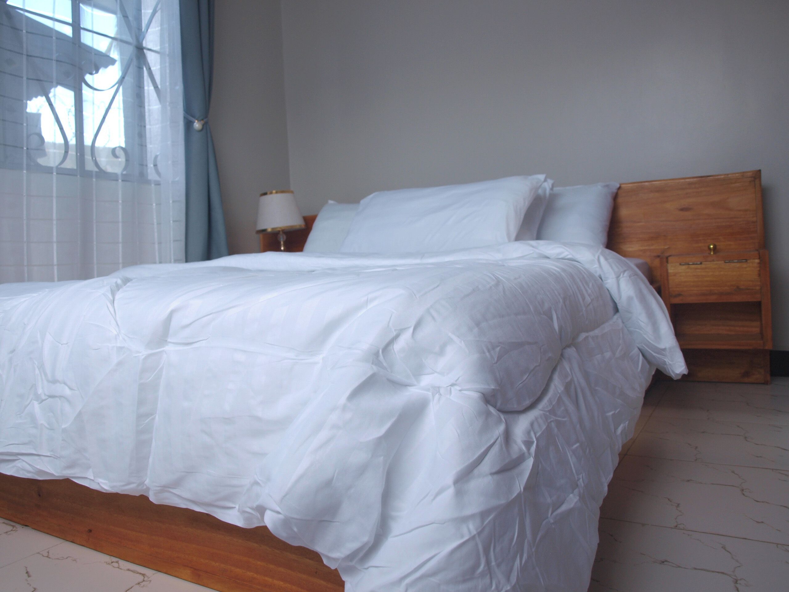 A bed with white bedding, a wooden headboard, a nightstand with a lamp, and a window with a curtain.
