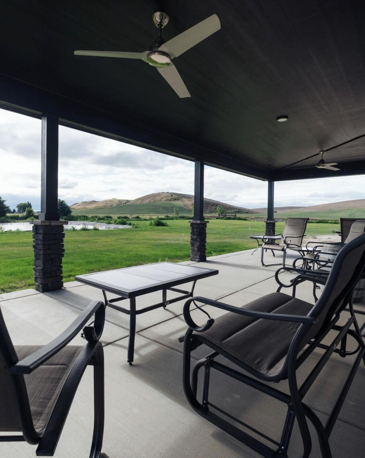 Une terrasse couverte avec une table et des chaises, un ventilateur de plafond et une vue sur un champ verdoyant et des collines.