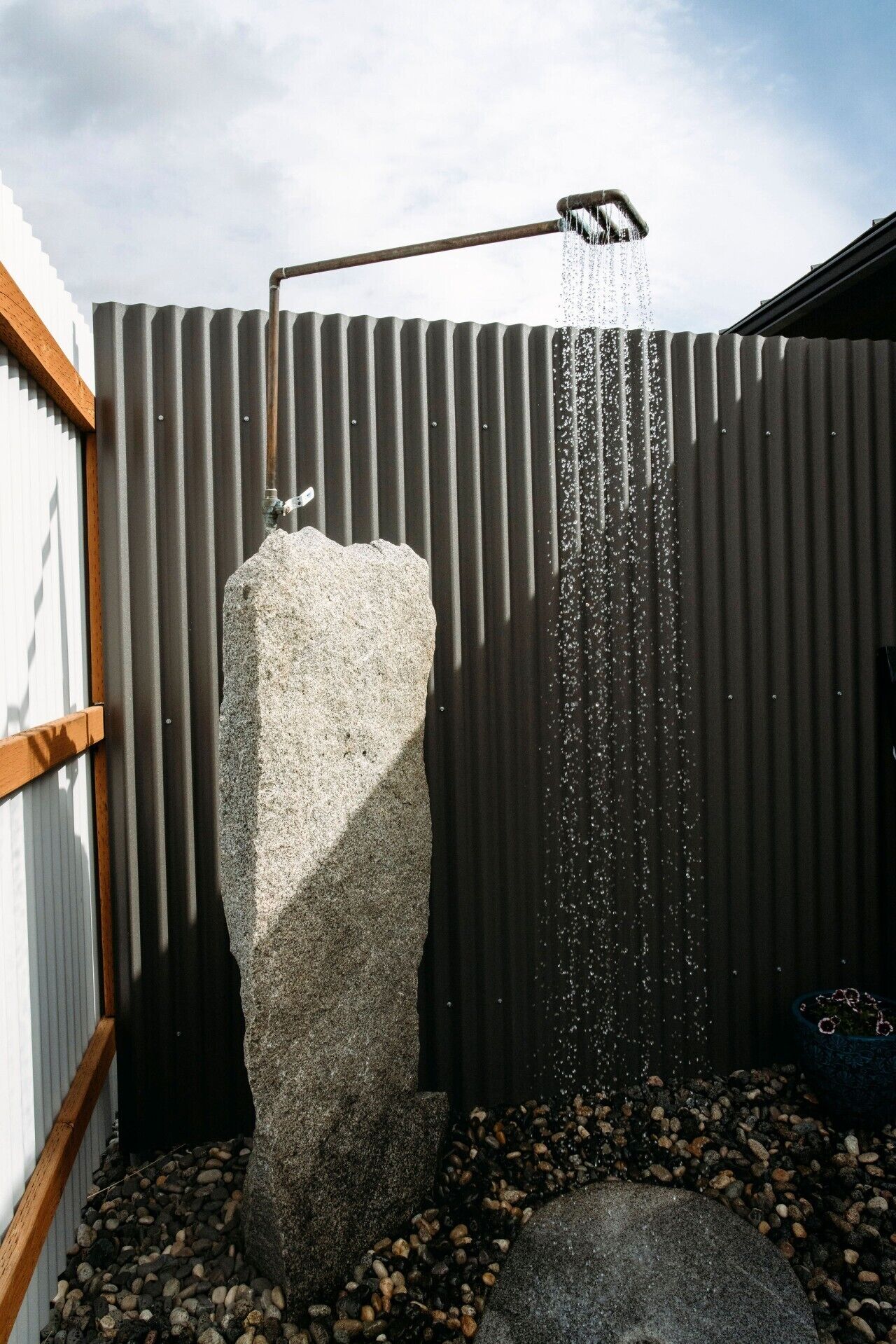 Un pommeau de douche fixé sur un mur en métal ondulé, l’eau s’écoulant le long d’une structure en pierre.