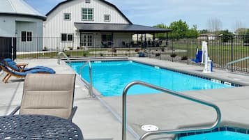 Une piscine dotée de chaises longues et d’un espace abrité, le tout entouré d’une clôture et bordé d’un bâtiment à toit en pignon.