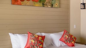 Cama com roupa de cama branca e duas almofadas vermelhas com estampa tropical, parede com pintura abstrata colorida e luminária pendente.