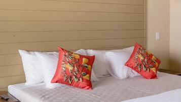 Una cama con sábanas blancas y dos almohadas rojas con estampado tropical, una pared con un cuadro abstracto colorido y una lámpara colgante.