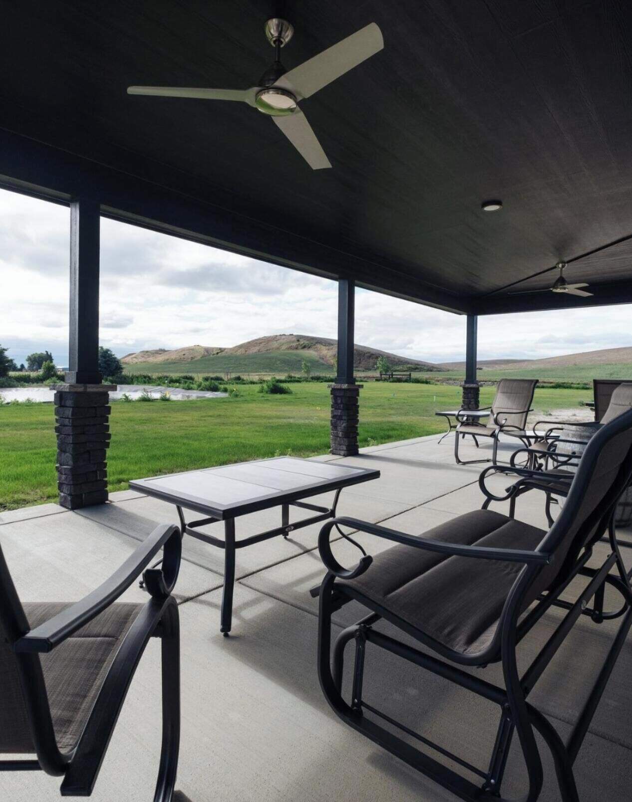 Une terrasse couverte avec une table et des chaises, un ventilateur de plafond et une vue sur un champ verdoyant et des collines.