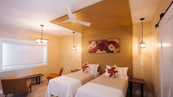 A hotel room with two beds, a desk, a chair, a ceiling fan, and a large window.