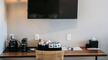 Een kamer met een wandgemonteerde flatscreen televisie, een houten bureau met een rieten stoel, een koffiezetapparaat en een prullenbak.