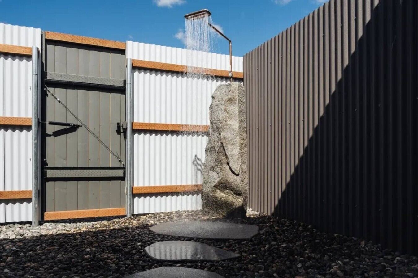 Une porte en métal agrémentée d’un élément décoratif à eau, un mur de pierre et un chemin recouvert de gravier.