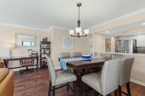 A dining area with a wooden table, chairs, a chandelier, and a kitchen in the background. - Seascape Towers 1025 (New Smyrna Beach)