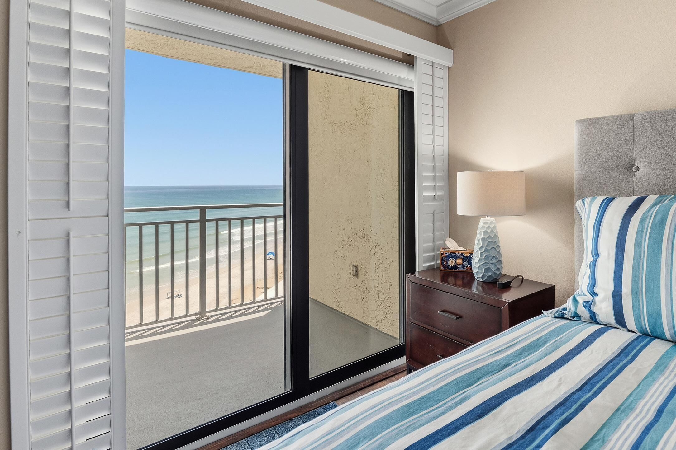A hotel room with a large sliding glass door leading to a beach view, a bed with striped bedding, a nightstand with a lamp, and a headboard.