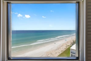 A beach view through a window, including the ocean, beach, and a building with a balcony. - Seascape Towers 1025 (New Smyrna Beach)