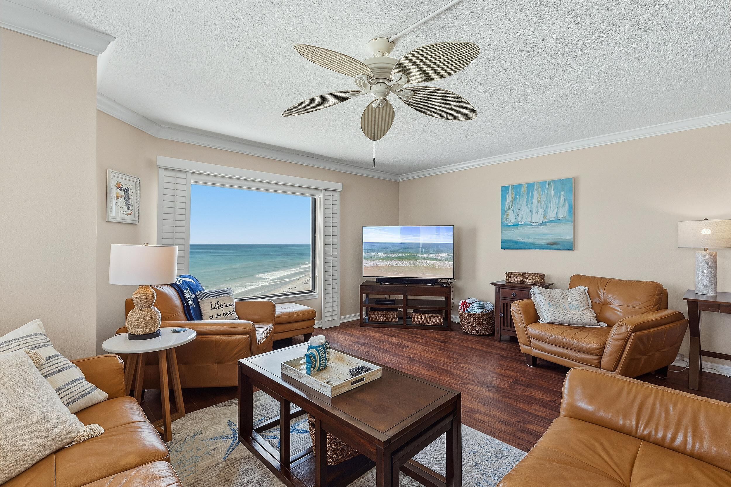 A living room with a view of the beach, leather furniture, a ceiling fan, and a television.