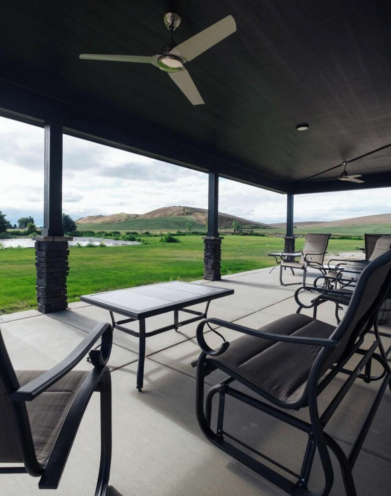 A covered patio with a table and chairs, overlooking a grassy field and hills.