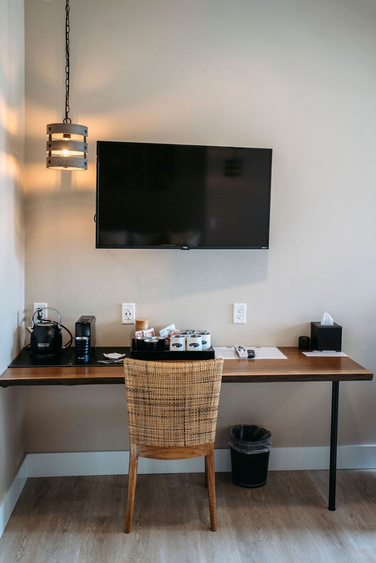 Une chambre dotée d’un téléviseur à écran plat fixé au mur, d’un bureau en bois avec une chaise en osier, d’une machine à café et d’une poubelle.