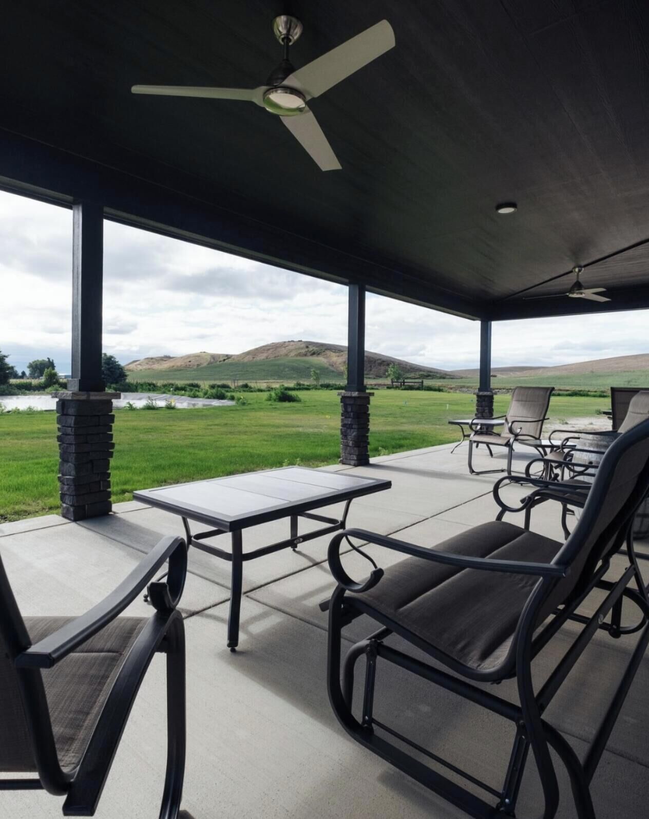 Une terrasse couverte avec une table et des chaises, un ventilateur de plafond et une vue sur un champ verdoyant et des collines.