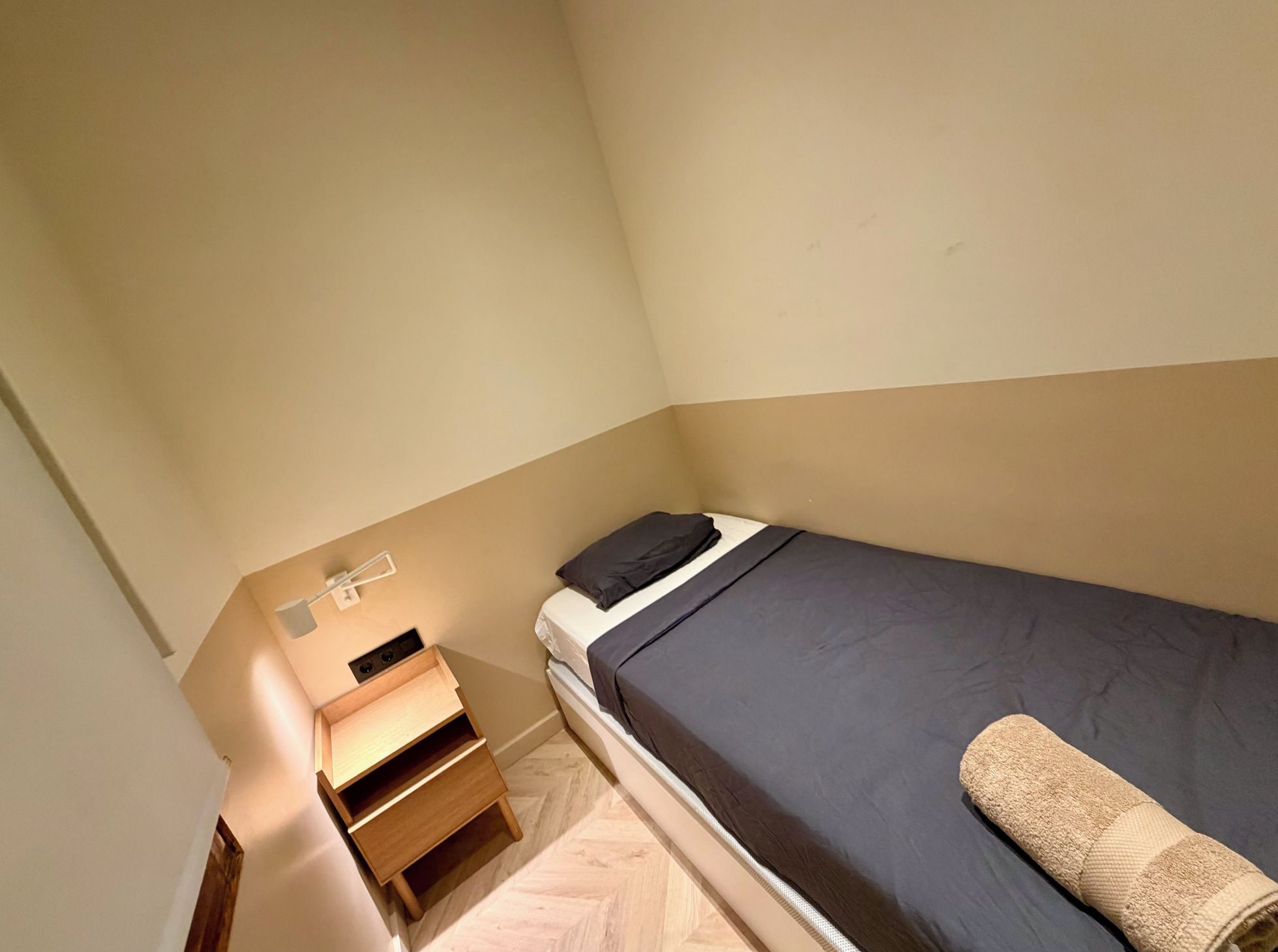 Ein kleines Zimmer mit einem Einzelbett, einem Nachttisch aus Holz und einer Lampe.