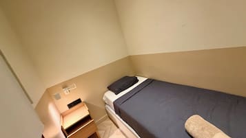 Ein kleines Zimmer mit einem Einzelbett, einem Nachttisch aus Holz und einer Lampe.