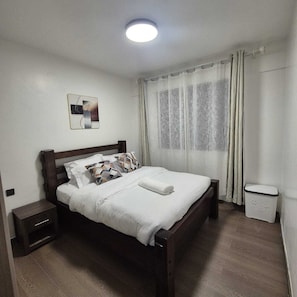 A bedroom with a wooden bed, white bedding, a nightstand, a wall-mounted picture, and a window with curtains. - Cozy Air BNB 2BR-Fast WiFi (Nairobi)