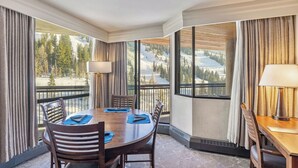 Quarto de hotel com uma grande mesa de jantar, cadeiras, um abajur e vista para uma paisagem montanhosa coberta de neve.