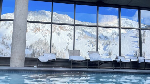 Área da piscina com espreguiçadeiras e vista para montanhas cobertas de neve através de grandes janelas.