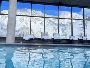 Un espace piscine avec des chaises longues et une vue sur les montagnes enneigées, visible à travers de grandes fenêtres.