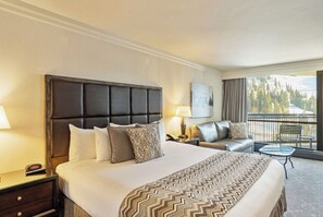 Habitación de hotel con una cama grande, un sofá, una mesita, una mesilla de noche y un balcón con vistas.