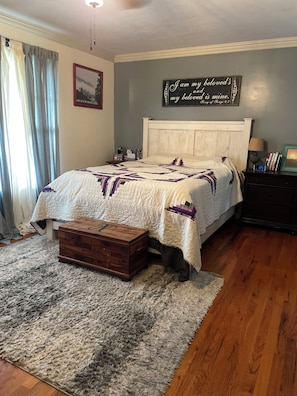 Une chambre à coucher comprenant un lit, une table de chevet, une commode en bois, un tapis et un mur orné d’un tableau encadré et d’une affiche.